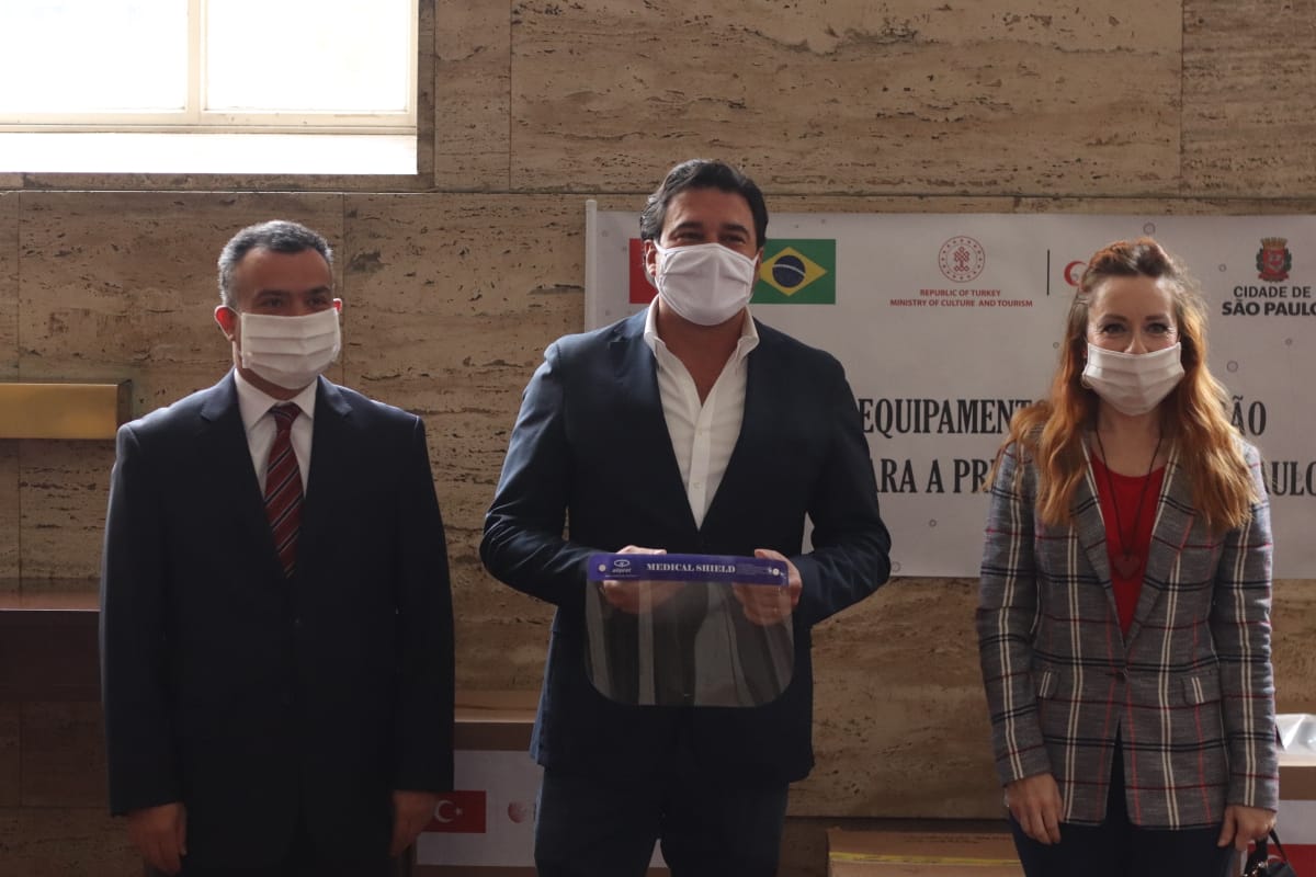Foto da cerimônia de entrega dos protetores faciais com o Cônsul-Geral da Turquia em São Paulo, Serkan Gedik, à direita, e o Secretário de Relações Internacionais da Prefeitura de São Paulo, Luiz Alvaro Salles Aguiar de Menezes, ao centro, segurando um protetor facial. 